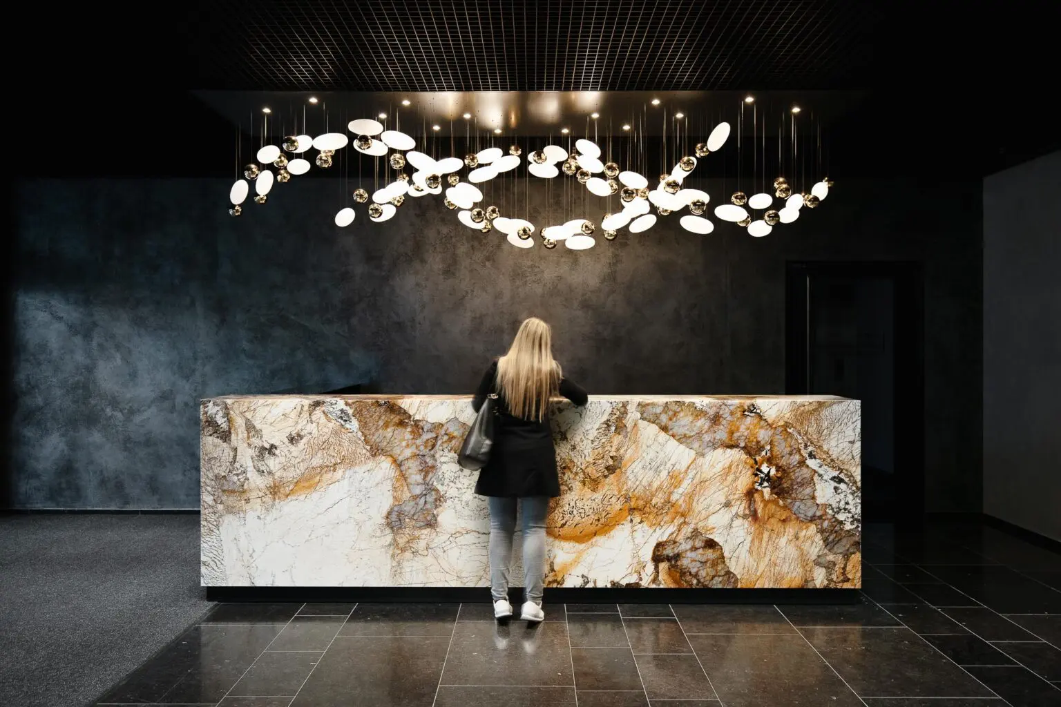 Designová lobby kanceláří s mramorovou recepcí, kruhovým lustrem a osobou s dlouhými vlasy.