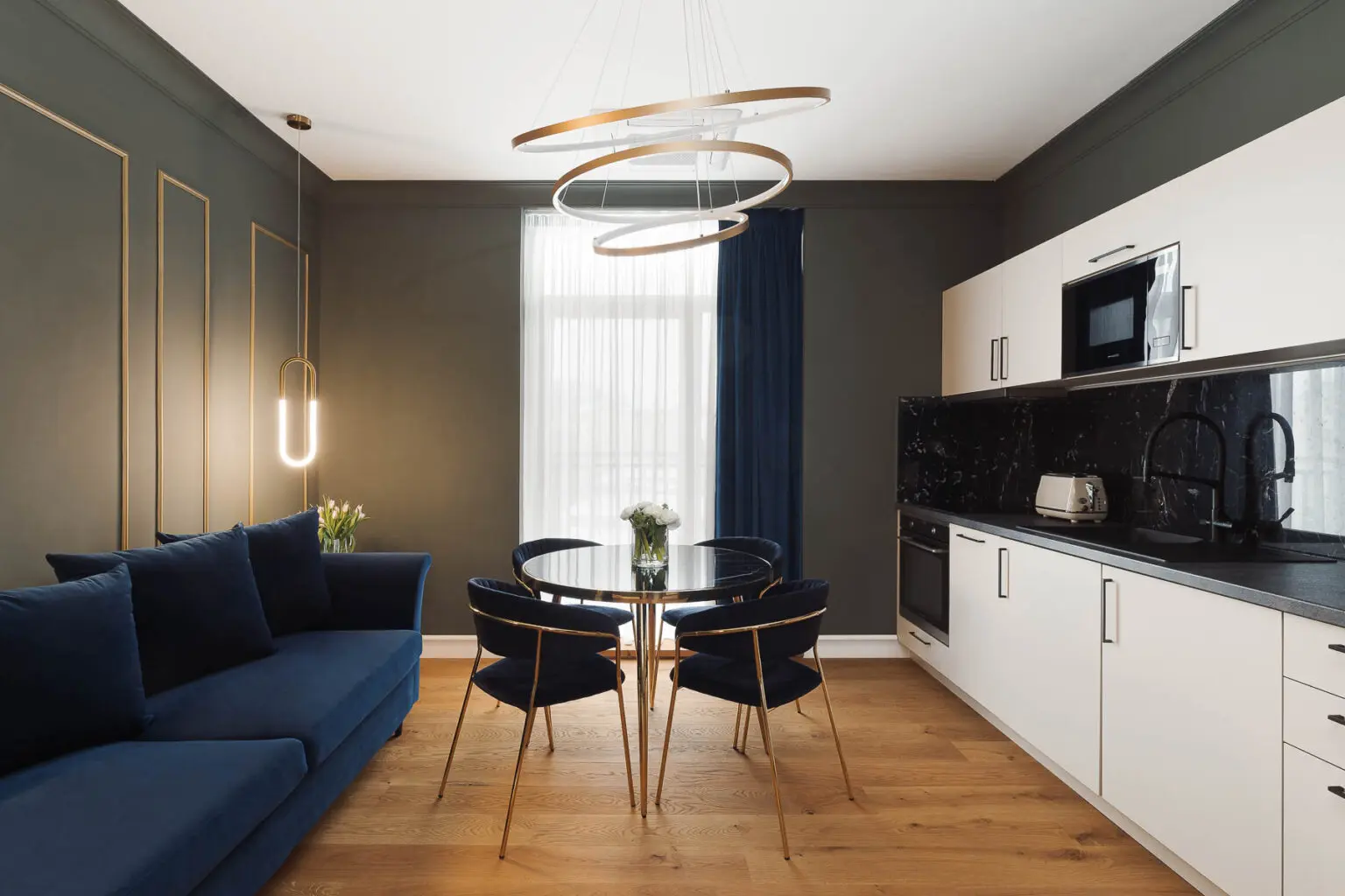 Moderní apartmán s bílou kuchyní a modrými luxusními židlemi. Ze stropu visí designový lustr.