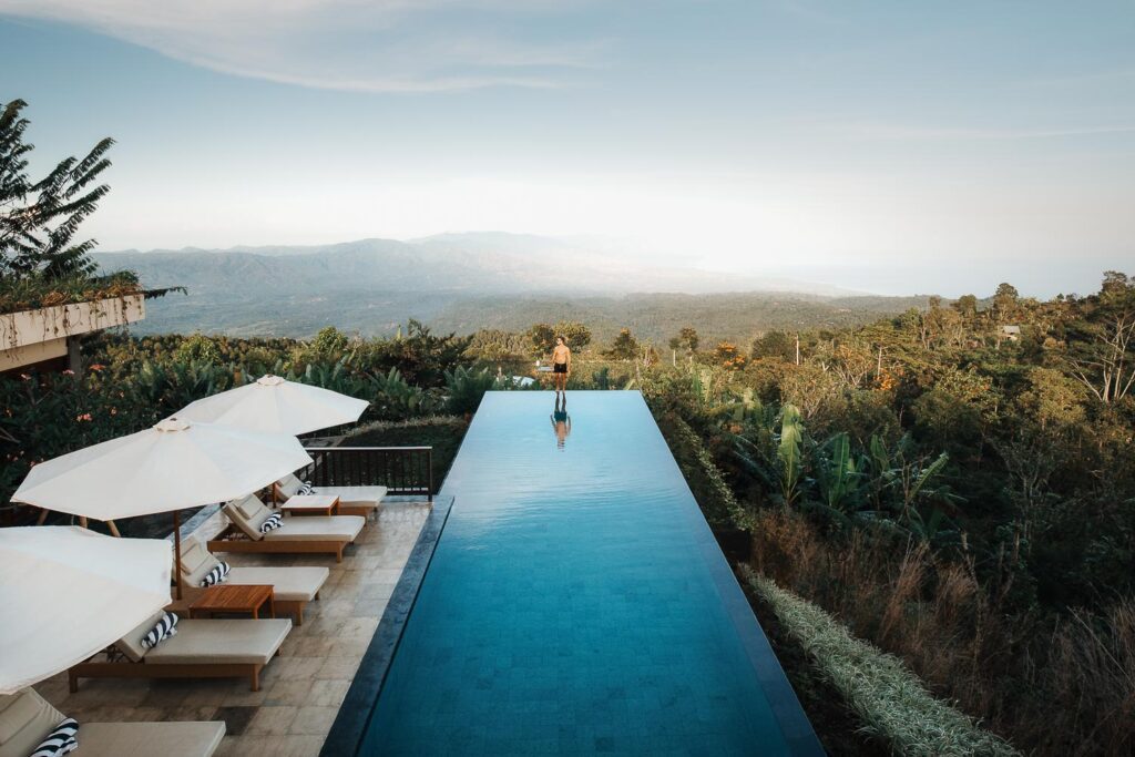 Infinity bazén na Bali s odrazem oblohy, lemovaný lehátky, s výhledem na hory a zeleň.