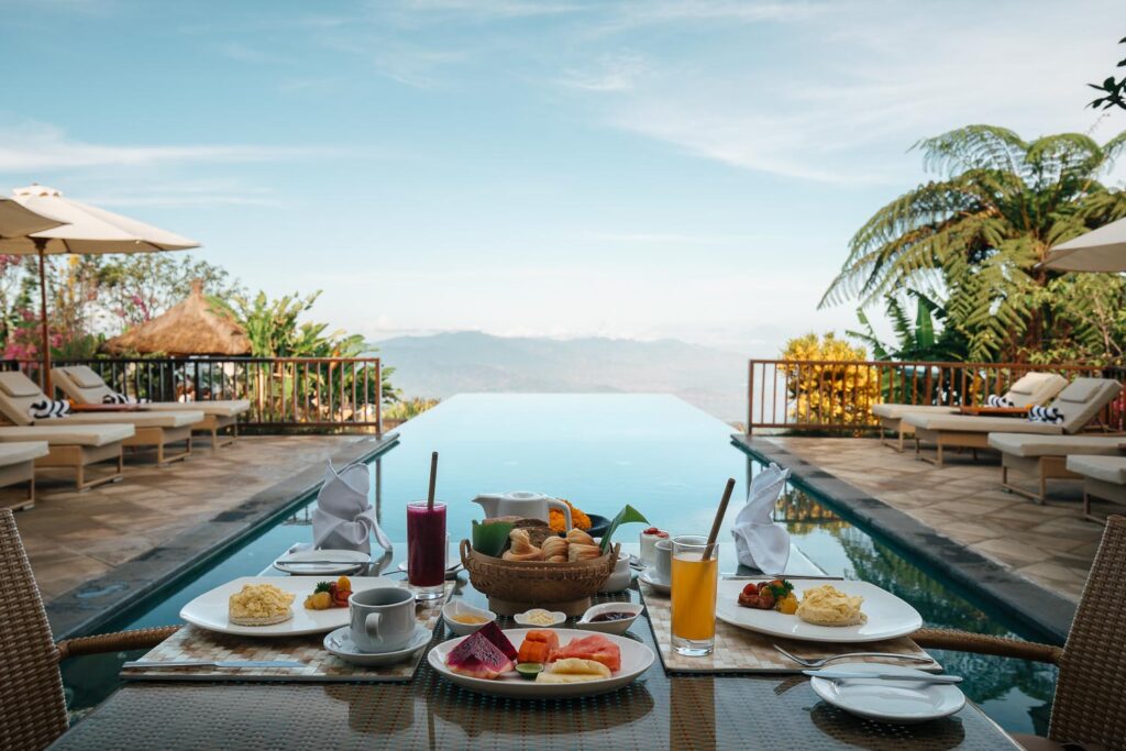 Výhled na nekonečný bazén luxusního hotelu na Bali se snídaní na stole a horami v pozadí.