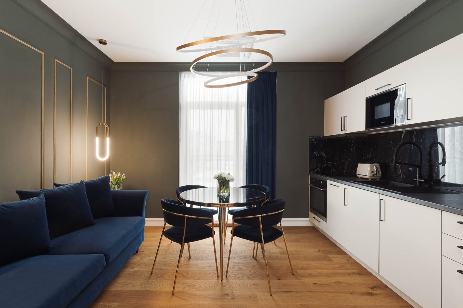 Moderní apartmán s bílou kuchyní a modrými luxusními židlemi. Ze stropu visí designový lustr.